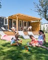 Camping La Plage Fleurie - Hébergements - Couple sur la terrasse de leur cottage et mère jouant avec ses enfants sur l\'herbe